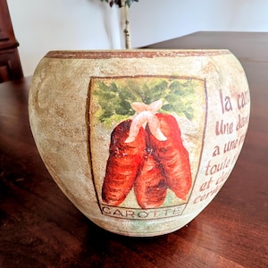 May include: A ceramic planter with a distressed, beige finish. It features a painted image of carrots with the French words "la carotte" and "carotte" on the front.