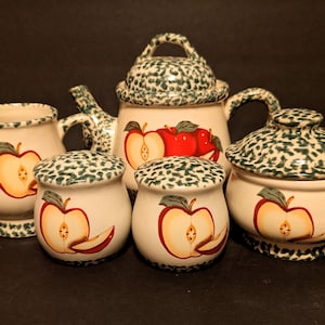May include: A ceramic apple-themed tea set with a teapot, creamer, sugar bowl, and two small jars. The set features a cream-colored base with red apple illustrations and green speckled accents. The teapot has a lid and handle.