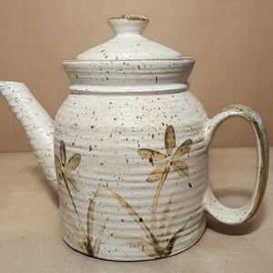 May include: A cream-colored ceramic teapot with a speckled finish and horizontal ridges. The body features hand-painted brown floral motifs with long stems and petals. It includes a matching lid with a knob handle, a curved spout, and a handle.