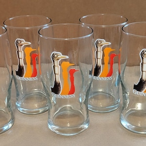 May include: Set of six clear glass pint glasses. Each glass features a colorful logo of a bird with the word "Guinness" below. The bird is black, white, orange, and red. The glasses are empty and sit on a light surface.