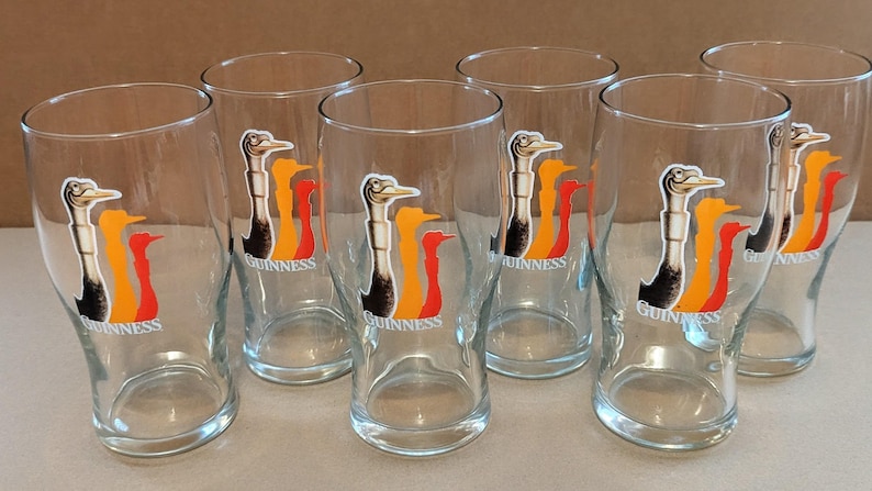 May include: Set of six clear glass pint glasses. Each glass features a colorful logo of a bird with the word "Guinness" below. The bird is black, white, orange, and red. The glasses are empty and sit on a light surface.