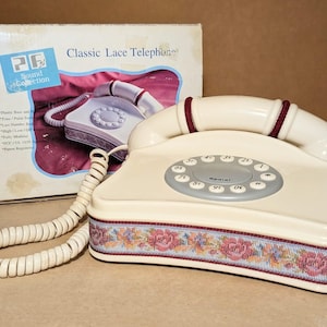 May include: A cream-colored corded telephone with a circular keypad and a handset featuring maroon accents. The base is decorated with a floral lace trim in shades of pink, blue, and gold. The original box labeled Classic Lace Telephone is included.