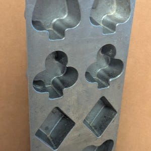 May include: A rectangular silver-colored metal baking mold featuring eight recessed cavities shaped like playing card suits. The mold includes two spade, two club, two diamond, and two heart-shaped indentations arranged in four rows with a small hole at the top.