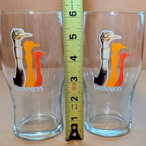 May include: Two clear pint glasses with the Guinness logo. Each glass features a colorful graphic of three stylized birds in shades of brown, orange, and red. The glasses are approximately 5 inches tall.