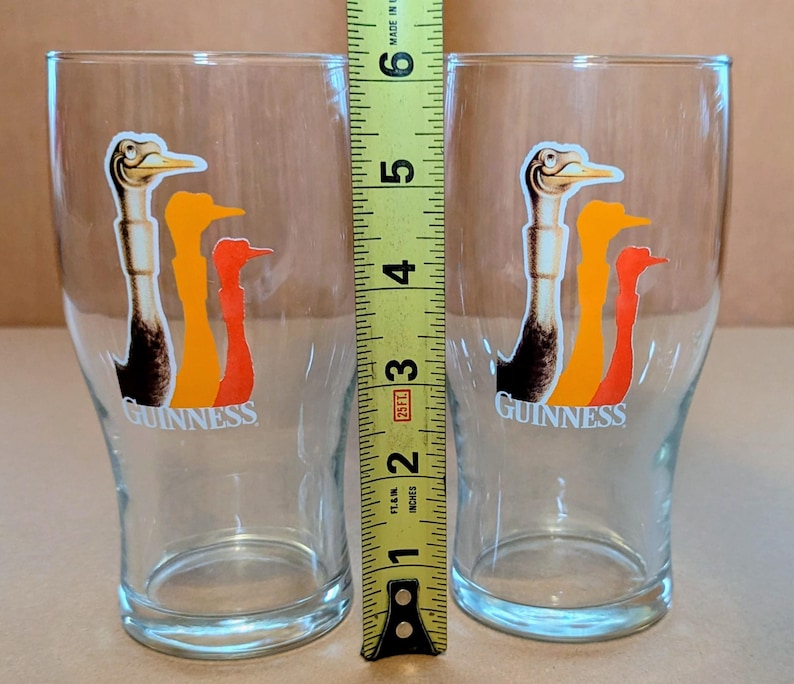 May include: Two clear pint glasses with the Guinness logo. Each glass features a colorful graphic of three stylized birds in shades of brown, orange, and red. The glasses are approximately 5 inches tall.