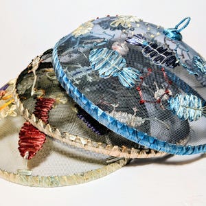 May include: Three decorative food covers with embroidered floral designs. The covers are made of a fine mesh material, with a blue, black, and tan color scheme. Each cover has a decorative trim around the edge and a loop for hanging.