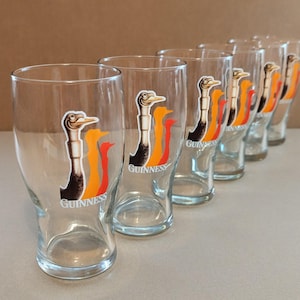 May include: Set of six clear pint glasses. Each glass features a colorful graphic of a bird with the word "Guinness" printed below. The glasses are arranged in a row, showcasing their design and clarity.