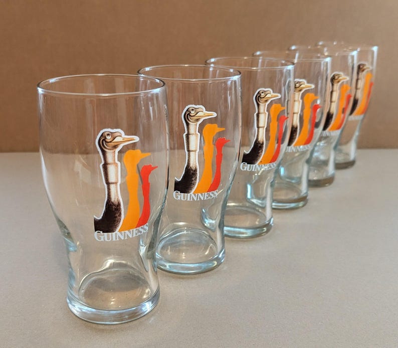 May include: Set of six clear pint glasses. Each glass features a colorful graphic of a bird with the word "Guinness" printed below. The glasses are arranged in a row, showcasing their design and clarity.