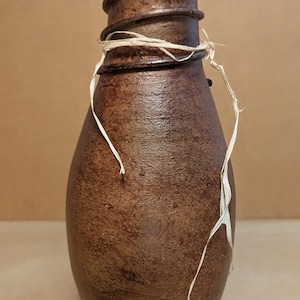 May include: A brown, textured, bottle-shaped object with a tapered neck and a wide base. The surface has a mottled, dark brown finish. A thin, light-colored raffia string is tied around the neck, and a spiral ridge wraps around the top.
