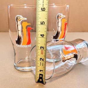 May include: Three clear pint glasses with the Guinness logo, featuring a stylized bird in black, orange, and red. The glasses are approximately 5 inches tall. One glass is lying on its side.