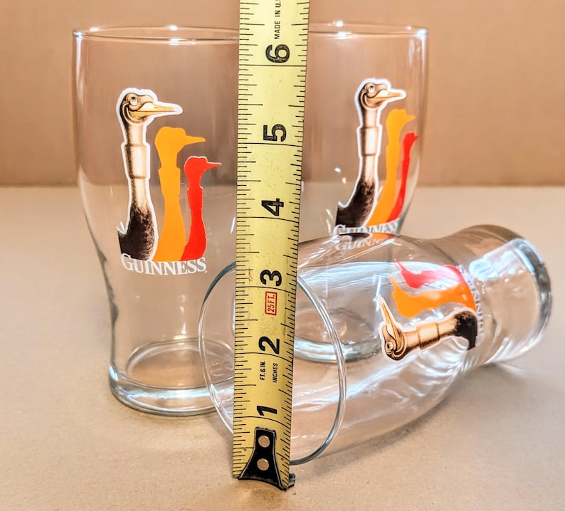 May include: Three clear pint glasses with the Guinness logo, featuring a stylized bird in black, orange, and red. The glasses are approximately 5 inches tall. One glass is lying on its side.