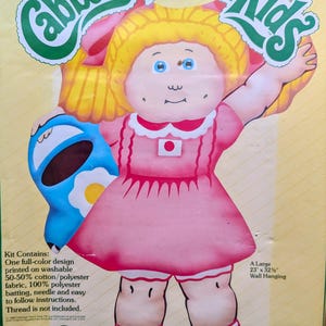 May include: A vintage Cabbage Patch Kids quilted wall hanging kit. The design features a doll in a pink dress holding a blue object. The kit includes a full-color design printed on washable fabric, batting, and instructions. The wall hanging measures 23" x 32".