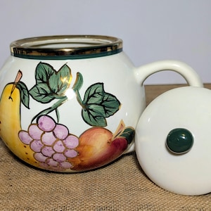 May include: White ceramic teapot with a gold rim and a painted fruit design. The teapot features a yellow pear, purple grapes, and a red apple. The lid is off, revealing a dark green knob. A classic kitchenware item.