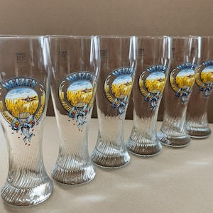 May include: A set of six tall, clear glass wheat beer glasses with a textured, swirling base. Each glass features a circular graphic depicting a golden wheat field, a small building, and a blue ribbon with the text WEIZEN printed above.