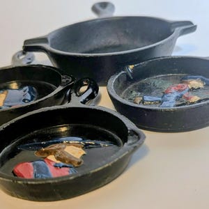May include: Set of four small, black cast iron pans, each with a handle or loop. The pans feature a relief of a person in a cowboy hat and work clothes. The pans vary in size and shape, with one larger oval pan.