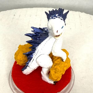 以下が含まれることがあります： 背中と頭に濃い青色のスパイクがある白い生き物のフィギュアで、質感のある黄色い物体を持っています。フィギュアは鮮やかな赤色のフェルトで覆われた円形の台座の上に立っており、縁には透明なプラスチックの枠が付いています。
