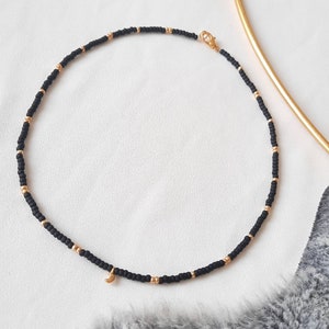 Könnte beinhalten: Eine zarte Halskette aus schwarzen Perlen mit einem goldenen Halbmond-Anhänger. Die Halskette besteht aus kleinen schwarzen Perlen und hat ein schlichtes, elegantes Design.