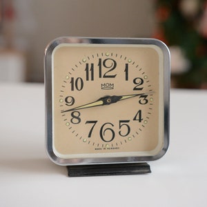 May include: A vintage silver alarm clock with a cream-coloured face. The clock has black numbers and hands, and the words "MOM" and "MADE IN HUNGARY" are printed on the face.