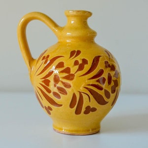 May include: A yellow ceramic pitcher with a handle and a bulbous body. The pitcher is decorated with a brown floral pattern. The pitcher is sitting on a white surface.