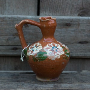 May include: A terracotta ceramic jug with a handle and spout. The jug is decorated with hand-painted floral designs in white, blue, and green. The jug is placed on a wooden surface.