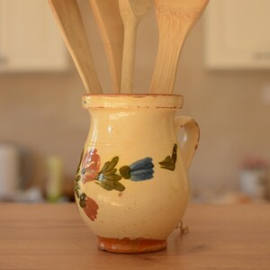 May include: A selection of wooden kitchen utensils, including spoons and spatulas, in a hand-painted ceramic jar. The jar has floral designs in red, blue, and green on a cream background. The utensils are made of light-coloured wood, and the jar has a handle.