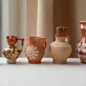 Op de afbeelding: Een verzameling van vijf kleine keramische kannen van verschillende groottes en vormen. De kannen hebben aardetinten, waaronder bruin, beige en crème, met handgeschilderde bloem- en bladpatronen in wit en groen op hun oppervlakken.