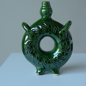May include: A circular green glazed ceramic vessel with a central opening, two side handles, and a small top spout. The surface features dark, intricate, hand-painted floral patterns and dots that contrast against the glossy, deep green finish.