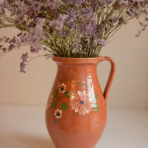 Pode incluir: Um jarro de terracota com uma única asa, apresentando motivos florais pintados à mão em branco e verde na parte frontal. O jarro está cheio com um grande ramo de flores de statice roxas secas, criando um contraste com a argila terrosa.