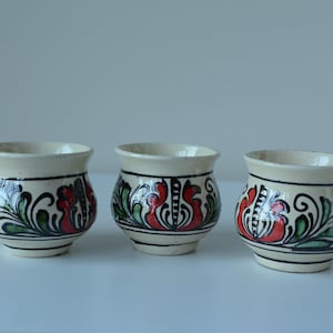 Può includere: Tre tazze in ceramica con base color crema, decorate con motivi floreali rossi, verdi e neri. Le tazze hanno una forma arrotondata, e una di esse ha un piccolo manico. Sono poste su una superficie bianca.