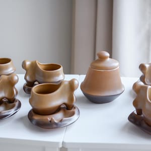 May include: A collection of brown ceramic teacups and saucers, along with a matching sugar bowl with a lid. The teacups have a unique, curved design. The set is displayed on a white surface.