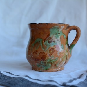 May include: A rustic ceramic mug with a handle. The mug has a brown base colour with green and orange abstract patterns. The mug is set against a white background with a striped cloth.