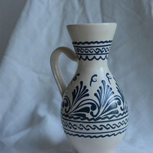 Peut inclure: Une cruche en céramique crème avec une anse et un dessus évasé. La cruche est décorée de motifs de vagues et floraux bleu foncé. La cruche est posée sur un tissu blanc.