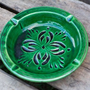 May include: A green ceramic ashtray with a black floral design. The ashtray has three indentations for cigarettes.