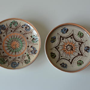 Puede incluir: Dos platos redondos de cerámica con intrincados patrones pintados a mano en tonos naranja, verde, azul y marrón. Los diseños incluyen motivos centrales en forma de estrella rodeados de elementos florales y líneas finas.