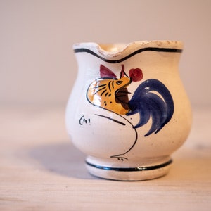 May include: White ceramic pitcher with a hand-painted rooster design. The rooster is yellow, red, and blue with a black outline. The pitcher has a rounded body and a small spout.
