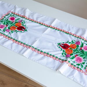 以下が含まれることがあります： 赤、オレンジ、ピンクのカラフルな花の模様が刺繍された白いテーブルランナー。花は緑の葉と赤と緑の縁で囲まれています。長方形のランナーは、おそらく綿でできています。