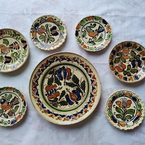 Pode incluir: Uma coleção de sete pratos redondos de cerâmica com fundo creme e motivos florais e de pássaros pintados à mão em tons de laranja, azul e verde. Os pratos variam em tamanho e textura da borda, alguns apresentam bordas recortadas.