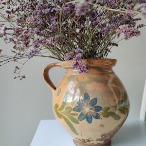 May include: A round, glazed ceramic pitcher with a beige base, featuring a hand-painted blue flower and green leaves on the front. The pitcher has a single side handle and is filled with a large bouquet of dried purple statice flowers.