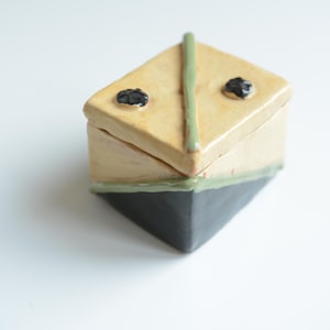 Può includere: Una scatola in ceramica fatta a mano con un design geometrico. La scatola è una combinazione di beige, nero e verde. Il coperchio ha due punti neri e una linea verde. La scatola misura circa 7.6 cm di larghezza e 7.6 cm di altezza.
