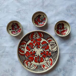 May include: A set of cream-colored ceramic tableware featuring hand-painted red and black floral motifs. The collection includes one large circular plate and three smaller matching bowls, each decorated with intricate botanical patterns and a bird.