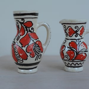 Może przedstawiać: Dwa białe dzbanki ceramiczne z czerwonymi i czarnymi wzorami kwiatowymi. Większy dzbanek ma szerszą podstawę i bardziej skomplikowany wzór. Mniejszy dzbanek ma węższą podstawę i prostszy wzór.