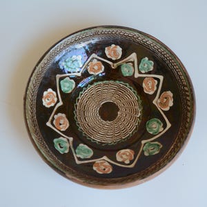 May include: A round ceramic bowl with a dark brown glazed interior featuring a central circular pattern of white lines. A white zigzag border surrounds the center, decorated with alternating light green and peach-colored floral motifs.