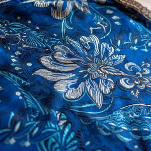 Può includere: Primo piano di un tessuto blu vibrante con un intricato motivo floreale in argento e azzurro. Il design presenta grandi fiori stilizzati e dettagli a spirale, creando un aspetto lussuoso e ornato. I bordi sono rifiniti con un bordo decorativo.
