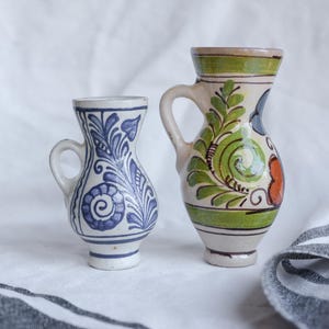 Può includere: Due piccoli brocchetti decorativi in ceramica. Il brocchetto a sinistra è bianco con disegni floreali blu. Il brocchetto a destra è color crema con motivi floreali verdi, rossi e neri. Entrambi hanno piccole maniglie.