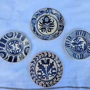 Könnte beinhalten: Vier runde Keramikteller mit handgemalten dunkelblauen Blumen- und geometrischen Mustern auf cremefarbenem Hintergrund. Jeder Teller zeigt ein einzigartiges Design, darunter zentrale botanische Motive, kunstvolle Ränder und gitterartige Details.