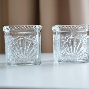 May include: Two clear glass candle holders with a seashell design. The holders are square and have a textured surface.