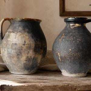 Può includere: Due antiche brocche in ceramica con manici. Le brocche sono grigio scuro con un aspetto usurato e strutturato e un bordo color crema. Sono appoggiate su una superficie di legno rustico, suggerendo un'estetica vintage o da fattoria. Le brocche sono probabilmente utilizzate per scopi decorativi.