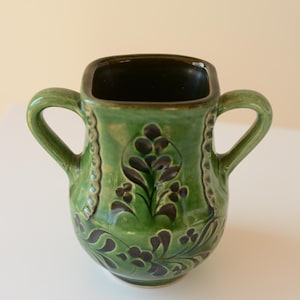 May include: Green ceramic vase with two handles and a square opening. The vase is decorated with dark green floral patterns and a decorative border. The interior is dark. A unique piece for home decor.