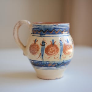 Può includere: Una tazza in ceramica con base color crema, decorata con bande orizzontali e linee ondulate dipinte a mano in blu. Il corpo presenta motivi floreali circolari arancioni ripetuti e pennellate blu verticali, completa di un manico laterale.