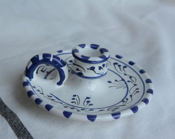 May include: A white ceramic candle holder with blue accents. The candle holder has a circular base with a raised center for the candle and a curved handle. The design includes blue dots and floral patterns.